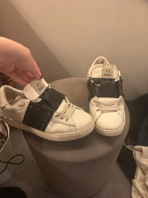 Vita sneakers från Valentino Garavani - Snygga vita sneakers från Valentino Garavani med svarta detaljer och nitar på sidan. Skorna har en låg profil, klassiska snören och en chunky sula. Märket syns tydligt på plösen och designen är riktigt clean men edgy.