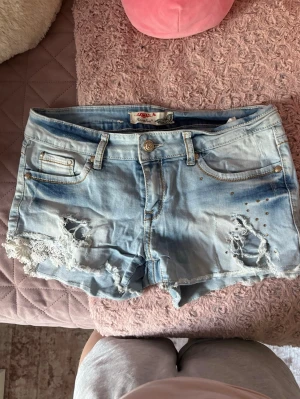 Snygga ljusblå jeansshorts - Säljer ett par super snygga ljusblå jeansshortsmed slitna detaljer, spets på ena benet och små nitar på framsidan. Perfekta för sommaren!!!🤍🤍🤍