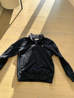 Marinblå tröja med dragkedja Polo Ralph Lauren - Mörkblå långärmad tröja från Polo Ralph Lauren med halv dragkedja och broderad logga på bröstet. Ribbstickade muddar och krage med röd och vit detalj. Perfekt för en avslappnad och stilren look.