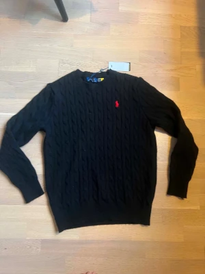 Svart kabelstickad tröja Polo Ralph Lauren - Kabelstickad svart tröja från Polo Ralph Lauren med rund halsringning och röd broderad logga på bröstet. Tröjan har långa ärmar och ribbade muddar vid ärmslut och nederkant. Perfekt för en stilren och klassisk look.