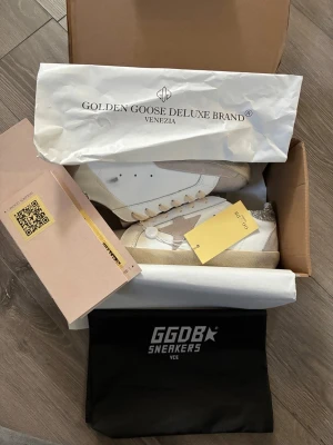 Golden Goose vita sneakers med stjärna - Golden Goose sneakers i vitt läder med beige mockadetaljer och den ikoniska stjärnan på sidan. Skorna har glitter på hälen och beige sula för en cool kontrast. Klassisk låg modell med snörning och GGDB-logga på sidan.