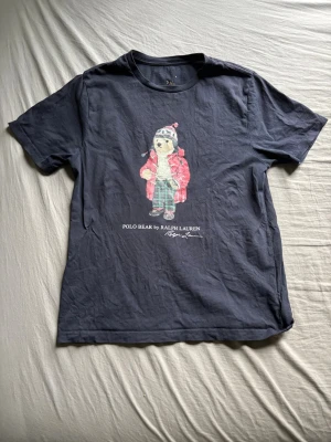 Mörkblå Polo Bear t-shirt Ralph Lauren - Snygg mörkblå t-shirt från Ralph Lauren med Polo Bear-tryck på bröstet. Klassisk passform med rund hals och korta ärmar. Tillverkad i mjuk bomull som känns skön mot huden. Perfekt för dig som gillar stilrena plagg med en twist.
