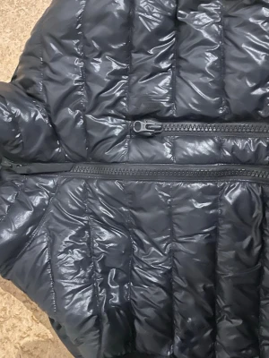 Svart dunväst med dragkedja - Snygg svart dunväst med quiltad design och tvåvägsdragkedja framtill. Västen har en glansig yta och är lätt vadderad för extra värme. Perfekt att slänga över en hoodie eller tröja för en cool lager-på-lager-look.