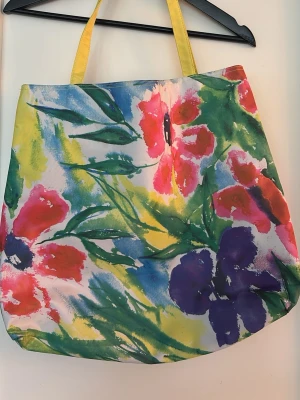 Färgglad shoppingväska med blommönster Lena Linderman - Stor shoppingväska med färgstarkt blommönster i rött, lila, grönt och gult. Väskan har gula axelband och är tillverkad i ett mjukt tygmaterial. Motiv av Lena Linderholm.