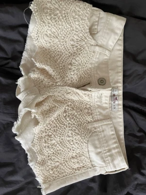 Vita spetsdetaljerade shorts Hollister - Säljer ett par vita shorts från Hollister med snygga spetsdetaljer på framsidan och råa kanter. Shortsen har klassisk femficksdesign, knapp och dragkedja framtill. Perfekta för varma dagar och ger en fräsch och trendig look.                                       OBS!!! Dock har knappen åkt av men om du vet hur man fixar det är shortsen så gott som nya!!