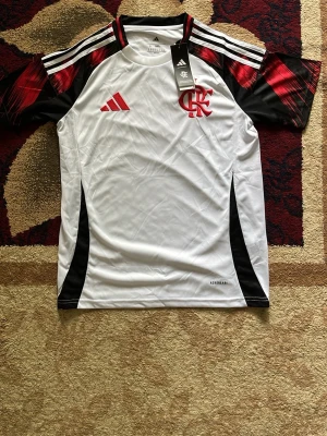 Fotbollströja  - Snygg vit matchtröja från Adidas med Flamengo-logga och röd Adidas-logga på bröstet. Svarta och röda detaljer på axlar och sidor, samt klassiska Adidas-ränder på axlarna. Tillverkad i lätt och ventilerande funktionsmaterial, perfekt för träning eller match.