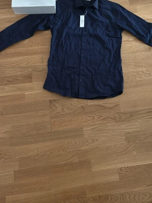 Mörkblå skjorta från John Henric - Stilren mörkblå skjorta från John Henric med klassisk krage och knäppning framtill. Skjortan är tillverkad i bomull och har lång ärm, perfekt för en clean och dressad look.