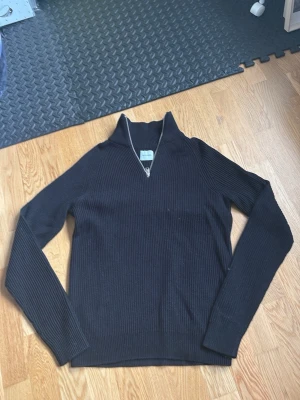 Svart stickad tröja med dragkedja - Snygg svart stickad tröja från Jack & Jones med halv dragkedja framtill och hög krage. Tröjan har ribbstickad struktur och långa ärmar, perfekt för lager på lager och en clean look.