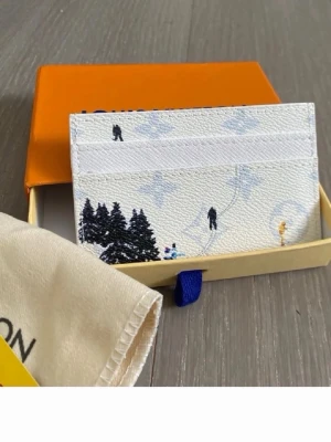 Louis Vuitton vit korthållare vintermotiv - Säljer en exklusiv vit korthållare från Louis Vuitton med vinterinspirerat motiv. Plånboken har diskreta LV-monogram, illustrationer av skidåkare och snötäckta granar. Tillverkad i tåligt läder och har flera kortfack. Perfekt accessoar för dig som gillar lyx och unik design.