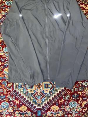 Svart nike Miller windbreaker - Snygg svart vindjacka från Nike med dragkedja framtill och klassisk vit Nike-logga på bröstet. Jackan har en minimalistisk design och är tillverkad i ett lätt, vindtätt material som passar perfekt för blåsiga dagar. Enkel och stilren look som funkar till det mesta.