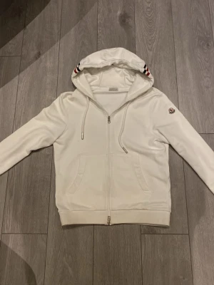 Vit hoodie från Moncler med dragkedja - Snygg vit hoodie från Moncler med dragkedja framtill och två fickor. Huva med dragsko och Monclers logga på ärmen. Detaljer i rött, vitt och blått på huva och ärmslut ger en sportig touch. Tillverkad i mjukt material som känns skönt mot huden.