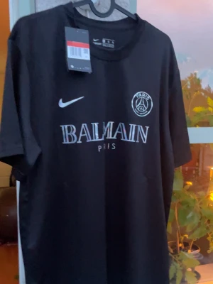 Svart PSG x Balmain t-shirt Nike - Snygg svart t-shirt från Nike i samarbete med PSG och Balmain. T-shirten har korta ärmar, rund hals och tryck med både Nike-logga och Paris Saint-Germain samt Balmain Paris på bröstet. Perfekt för dig som gillar streetwear och sportig stil.