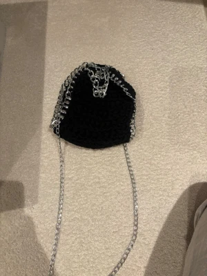 Svart stickad handväska med kedja - Cool svart handväska i grovstickat tyg med silverfärgad kedja som axelrem och detaljer runt kanterna. Väskan har en mjuk, påsliknande form och är perfekt för dig som gillar en edgy och trendig look.