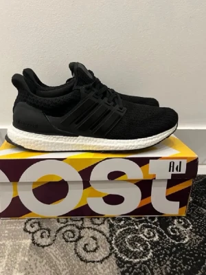 Adidas Ultra Boost svarta sneakers - Svarta Adidas Ultra Boost sneakers med stickad ovandel och vita Boost-sulor. Klassisk design med tre svarta ränder på sidan och svart hälkappa. Sköna och lätta med sportig känsla, perfekta för dig som gillar streetwear och komfort.