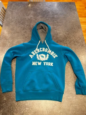Blå hoodie från Abercrombie - Snygg blå hoodie från Abercrombie med vit broderad text 'Abercrombie New York' och logga på bröstet. Hoodien har en stor ficka framtill, vita dragsnören och grått innerfoder i huvan. Perfekt för en avslappnad stil.