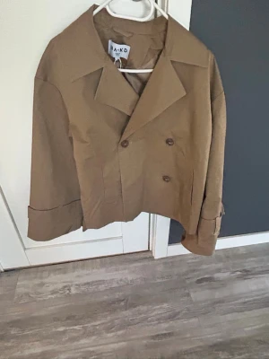 Kort beige trenchcoat från NA-KD - Snygg kort trenchcoat i beige från NA-KD med dubbelknäppning och bred krage. Jackan har uppvikta ärmslut och en rak, boxig passform. Perfekt för dig som vill ha en trendig och stilren look. Tillverkad i ett lätt material som passar bra till vår och höst. Jackan är i storlek 32 men passar lätt som 34 för den är översized.
