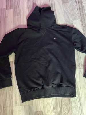 Svart hoodie från Tommy Hilfiger - Svart hoodie från Tommy Hilfiger. Använd Max 5 gånger.  med liten logga på bröstet.  Klassisk modell med huva, ribbade muddar vid ärmslut och nederkant. Tillverkad i mjukt bomullsmaterial som känns skönt mot huden. Perfekt för en chill och stilren look.