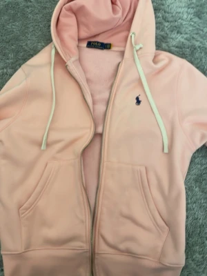 rosa hoodie från Polo Ralph Lauren - rosa hoodie från Polo Ralph Lauren med dragkedja, vit dragsko i huvan och klassisk blå logga broderad på bröstet. Hoodien har två fickor framtill och är tillverkad i mjukt material som känns riktigt skönt mot huden.