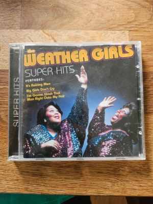 Weather Girls Super Hits CD - CD-album med Weather Girls 'Super Hits' i plastfodral. Omslaget har färgstark design med glitterklädda artister och låtar som 'It's Raining Men' och 'Big Girls Don't Cry'. Perfekt för dig som gillar klassisk disco och soul.