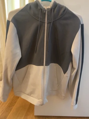 Gråvit hoodie med dragkedja från boss - Snygg hoodie i grått och vitt med dragkedja framtill och justerbar huva. Hoodien har en modern tvåfärgad design och är tillverkad i mjukt sweatshirtmaterial. Perfekt för en avslappnad och trendig stil.