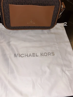 Brun axelväska från Michael Kors - Snygg axelväska från Michael Kors i brunt konstläder med logomönster och en slät ljusbrun ficka framtill. Väskan har guldfärgade detaljer och justerbar axelrem. Perfekt storlek för det viktigaste och ger en trendig look till din outfit.
