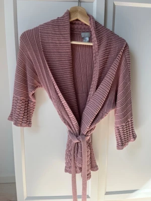 Rosa stickad kofta från H&M - Säljer en mysig stickad kofta i dammigt rosa från H&M. Koftan har trekvartsärmar, bred krage och ett matchande knytband i midjan. Ärmsluten och nederkanten har strukturstickade detaljer som ger extra stilpoäng. Perfekt för lager på lager och chill dagar.