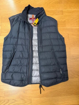Svart dunväst från Parajumpers - Snygg svart dunväst från Parajumpers med quiltad design och hög krage. Västen har två dragkedjeförsedda fickor framtill och en gul detalj vid halsen. Insidan är ljusgrå och materialet är lätt och smidigt, perfekt för lager-på-lager.