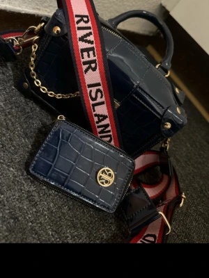 Mörkblå handväska från River Island - Snygg mörkblå handväska i krokopräglat konstläder från River Island. Väskan har guldfärgade detaljer, bred axelrem med logga i rött och vitt samt matchande liten plånbok. Boxig form och kedjedetalj ger en trendig look. Priset kan diskuteras 