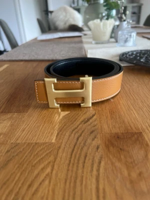 Skärp med guldfärgat H-spänne - Snyggt brunt orange skärp med guldfärgat H-spänne. Bältet har vita kontrastsömmar och är gjort i exklusivt skinn. Perfekt accessoar för att lyfta vilken outfit som helst. Hör av er vid intresse. Priset går att diskutera.
