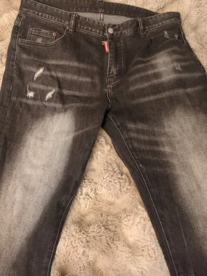 Svarta Dsquared2 jeans med slitningar - Säljer ett par svarta Dsquared2 jeans med coola slitningar och tvättade detaljer. Jeansen har klassisk femficksmodell, knappgylf och en stor 'CIAO' patch bak i midjan. Materialet är denim i bomull och passformen är rak.