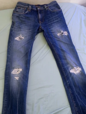 Blå slitna jeans från Nudie Jeans - Säljer ett par blå jeans från Nudie Jeans med slitna detaljer och hål på både lår och knän. Jeansen har klassisk femficksmodell, orangea sömmar och är tillverkade i denim. Perfekta för en avslappnad och trendig look.