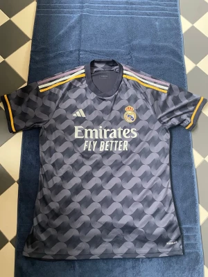 Real Madrid matchtröja mörkgrå - Snygg mörkgrå Real Madrid matchtröja från Adidas med vågigt mönster, gula detaljer och klubbmärke på bröstet. Tryck med Emirates Fly Better på framsidan och nummer 10 i guld på ryggen. Tillverkad i lätt och ventilerande funktionsmaterial. Tröjan var för stor så den har bara användts en gång. 