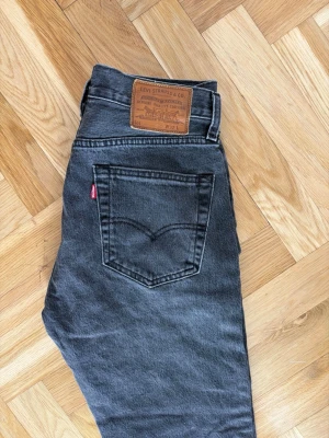Svarta Levi's raka jeans - Säljer ett par svarta raka jeans från Levi's med klassisk femficksdesign och läderpatch bak i midjan. Jeansen har en vintagekänsla och är tillverkade i kraftig denim med en diskret tvättad look. Perfekta för en avslappnad och stilren outfit.