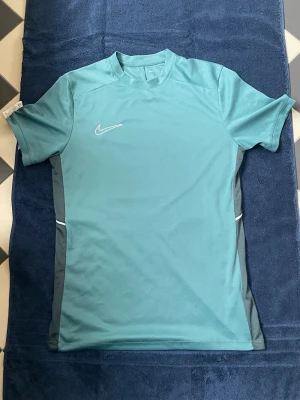 Turkos Nike träning/gym tröja - Snygg turkos träningströja från Nike med korta ärmar och rund hals. Tröjan har mörkare sidopaneler och är tillverkad i ett lätt, snabbtorkande funktionsmaterial som passar perfekt till träning, gym och sportaktiviteter. Diskret Nike-logga på bröstet. Använd ett fåtal gånger (2-4 gånger)