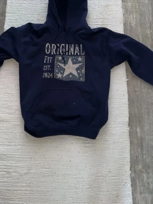 Mörkblå hoodie med tryck - Snygg mörkblå hoodie med huva och magficka. Framsidan har ett stort tryck med texten 'ORIGINAL FIT EST. 2024' och en stjärna omgiven av små stjärnor. Perfekt för en avslappnad och cool stil.