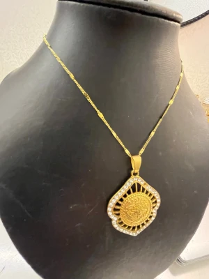 Guldigt halsband med stor medaljong - Unikt halsband i guldfärg med en stor, dekorativ medaljong. Medaljongen har ett detaljerat mönster i mitten och är kantad av små pärlor som ger extra bling. Kedjan är tunn och elegant, vilket gör att hänget verkligen står ut. Perfekt för dig som gillar statement-smycken.