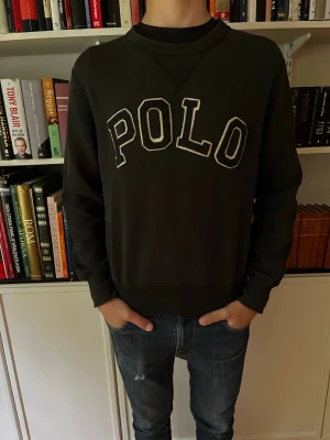 Svart sweatshirt från Polo Ralph Lauren - Cool svart sweatshirt från Polo Ralph Lauren med stor vit POLO-text på bröstet. Tröjan har rund halsringning, ribbade muddar och avslappnad passform. Perfekt till jeans för en casual look.