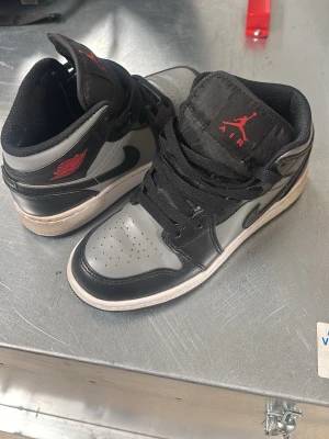 Air Jordan 1 Mid svart/grå - Snygga Air Jordan 1 Mid sneakers i svart och grått läder med röda detaljer på plösen och hälen. Klassisk siluett med svart swoosh, perforerad tåbox och vadderad krage. Skorna har svarta skosnören och vit sula.