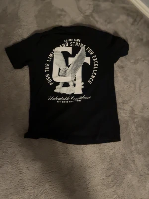Svart t-shirt med tryck och vinge - Snygg svart t-shirt med stort vitt tryck på ryggen och mindre tryck fram. Trycket har bokstäver och en vinge, samt texten 'Push the limit and strive for excellence'. Klassisk passform och rund hals. Perfekt för dig som gillar streetwear.