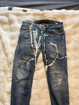 Blå jeans från Dondup  med färgstänk och slitningar - Säljer ett par blå jeans med coola vita färgstänk och slitna detaljer på både fram- och baksida. Jeansen har en smal passform och klassisk femficksdesign. 