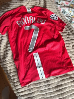 Manchester United Ronaldo tröja - Röd Manchester United matchtröja med Ronaldo och nummer 7 på ryggen. Tröjan har Champions League-märke på ärmen, AIG som sponsor på bröstet och klubbmärke i gult och rött. Tillverkad i lätt och ventilerande material, perfekt för fotbollsträning eller match.