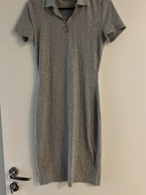 Grå ribbad klänning från FB Sister - Grå ribbad kortklänning från FB Sister med korta ärmar och krage. Klänningen har en dragkedja framtill med rund metallring som detalj. Perfekt för en avslappnad och trendig look.