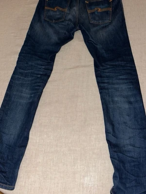 Mörkblå jeans från Nudie Jeans - Säljer ett par mörkblå jeans från Nudie Jeans med klassisk femficksmodell och snygga slitningar framtill. Jeansen har raka ben och kontrastsömmar, samt Nudie Jeans signaturbrodyr på bakfickorna. Perfekta för en avslappnad och trendig look.