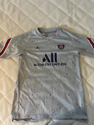 Grå PSG matchtröja Jordan x Nike - Snygg grå PSG matchtröja med svarta prickar, blå logga och texten 'Accor Live Limitless' på bröstet. Röd, vit och svart detalj på ärmarna samt PSG-märke och Jordan-logga. Tillverkad i lätt och ventilerande material, perfekt för fotboll eller träning.