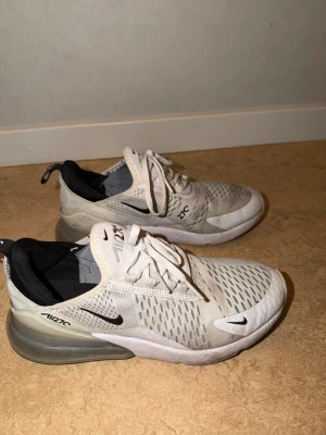 Nike Air Max 270 vita sneakers - Nike Air Max 270 sneakers i vitt med svarta detaljer och synlig Air Max-sula. Ovandelen är i mesh för bra andningsförmåga och har klassisk snörning. Snygg och sportig design med logga på sidan och häl. Perfekta för en streetwear-look.
