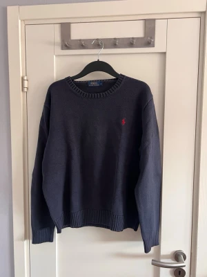 Mörkblå stickad tröja Polo Ralph Lauren - Klassisk mörkblå stickad tröja från Polo Ralph Lauren med rund halsringning och ribbade muddar. Tröjan har den ikoniska röda logotypen broderad på bröstet. Perfekt för lager på lager och enkel att matcha med olika stilar.