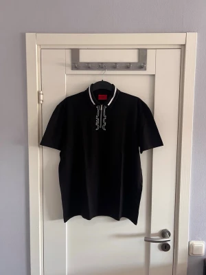 Svart Hugo Boss t-shirt med tryck - Snygg svart t-shirt från Hugo Boss med vit logga och tryck på kragen. T-shirten har korta ärmar och klassisk passform. Perfekt till jeans eller shorts för en clean och stilren look.