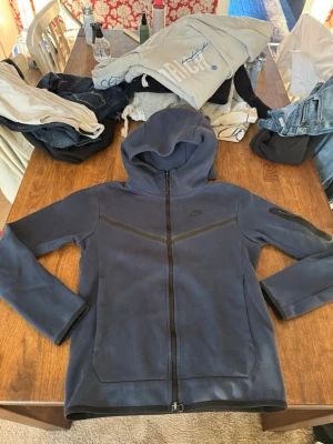 Mörkblå Nike hoodie och byxor med dragkedja - Snygg mörkblå hoodie från Nike med hel dragkedja framtill och huva. Hoodien har svarta detaljer längs bröstet och ärmarna samt diskret Nike-logga på bröstet. Tillverkad i mjukt och bekvämt material, perfekt för en avslappnad stil.