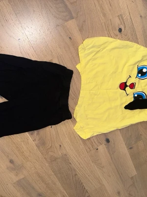 Gult Pikachu-inspirerat set - Sött set med gul body och svarta byxor. Bodyn har ett stort Pikachu-liknande ansikte med blå ögon och röda kinder framtill samt svarta öron med knyt på axlarna. Tillverkad i mjuk bomull, perfekt för dig som gillar Pokémon och vill ha något lekfullt.
