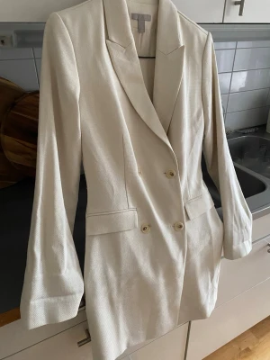 Kavaj  - Stilren beige  kavaj med dubbelknäppning och två fickor framtill. Kavajen har klassisk krage och är tillverkad i ett strukturerat tyg som ger en exklusiv känsla. Perfekt för dig som vill ha en clean och modern look. Oanvänd